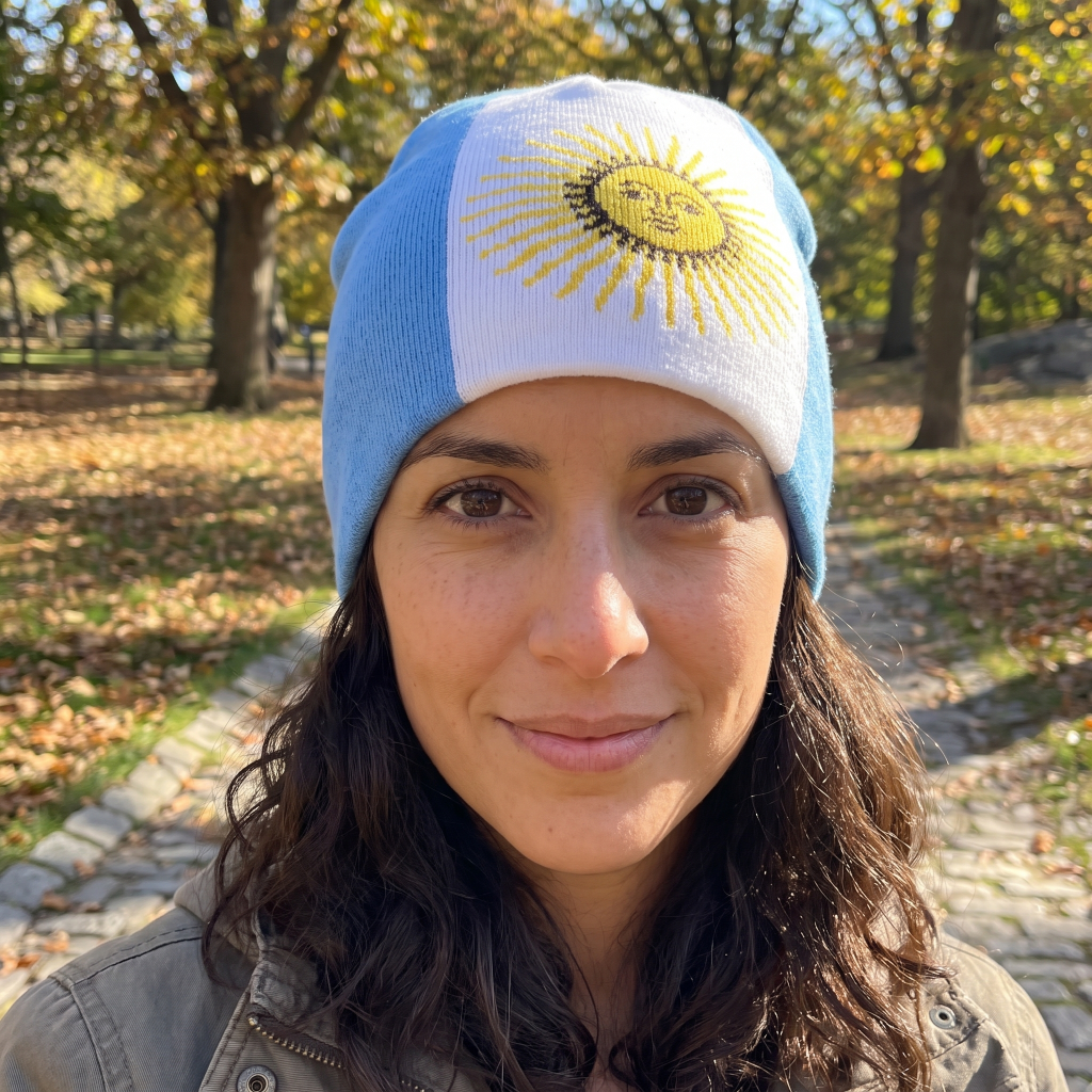A woman wearing a light blue and white knit beanie with a yellow embroidered sun emblem, photographed outdoors in a park with trees and fallen leaves in the background.