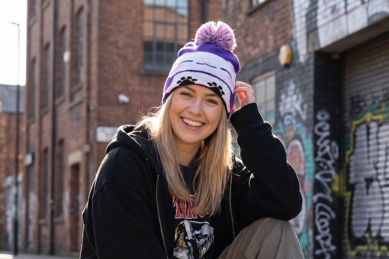 Person wearing a black hoodie and colorful beanie squatting on a street with graffiti in the background