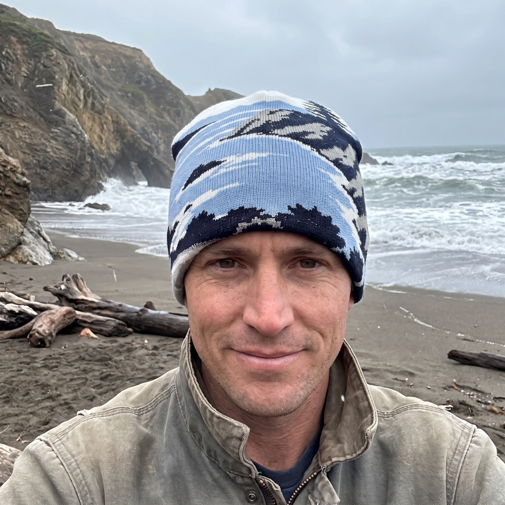 A man wearing a blue and navy patterned knit beanie hat and a beige jacket, photographed on a beach with ocean waves and rocky cliffs in the background.