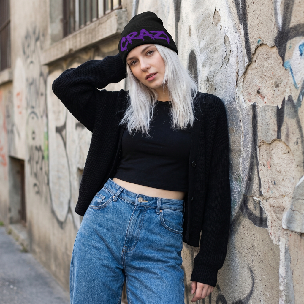 Person wearing a black beanie with 'CRAZY' text, black crop top, and blue jeans against a graffiti-covered wall.