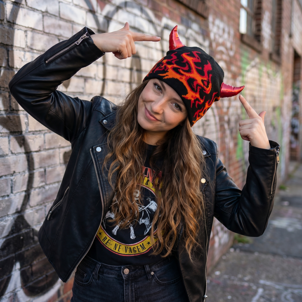 Person wearing a black leather jacket and a colorful flame-patterned hat with red horns, standing against a brick wall.