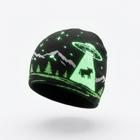 A black knit beanie hat featuring a glowing green and gray graphic of a UFO, mountain peaks, pine trees, and stars, shown against a light gray background.