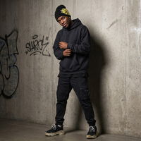 Person wearing a dark hoodie and pants leaning against a graffiti-covered wall.