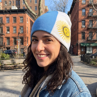 A smiling woman outdoors wearing a knit beanie with a light blue and white vertical split and a yellow sun emblem on the white side, resembling the Argentina flag. Brick apartment buildings and a street scene are visible in the background.