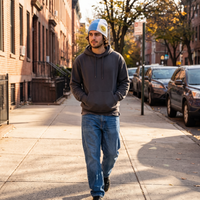 A man walking on a city sidewalk wearing a dark gray hoodie, blue jeans, and a light blue and yellow beanie, with brick buildings and parked cars in the background.