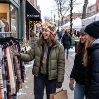 A woman in an olive green puffer jacket and a patterned beanie smiles while browsing scarves on a display rack at an outdoor winter street market, with two other shoppers beside her.