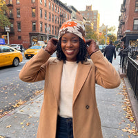 A woman smiling on a city sidewalk wearing a tan coat, a cream sweater, dark jeans, and a camouflage beanie with orange accents. Brick apartment buildings and yellow taxis are visible in the background.