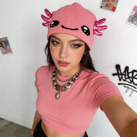 Person wearing a pink beanie with axolotl design and matching pink top, standing against a white wall.