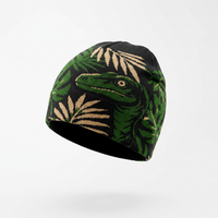 A knit beanie hat featuring a green dinosaur head and palm leaves on a black background, shown floating against a light gray studio backdrop.