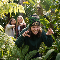 A woman in a dark green hoodie and a green beanie with a leaf pattern smiles while crouching among lush green ferns, with two other women playfully pointing in the background.