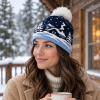 A woman wearing a navy and light blue knit beanie with a white pom-pom and winter landscape pattern, holding a mug in front of a cozy cabin window during winter snowfall.