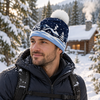 A man wearing a navy blue knit beanie with a white pom-pom and a snowy mountain pattern, standing outdoors in a snowy mountain setting with a cabin in the background.