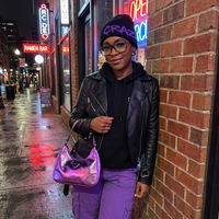 Person wearing a black leather jacket, purple pants, and a neon beanie leaning against a brick wall with neon signs in the background.