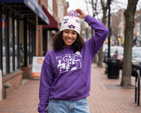 Person wearing a purple hoodie with a logo and a white beanie with purple flowers, standing on a sidewalk.