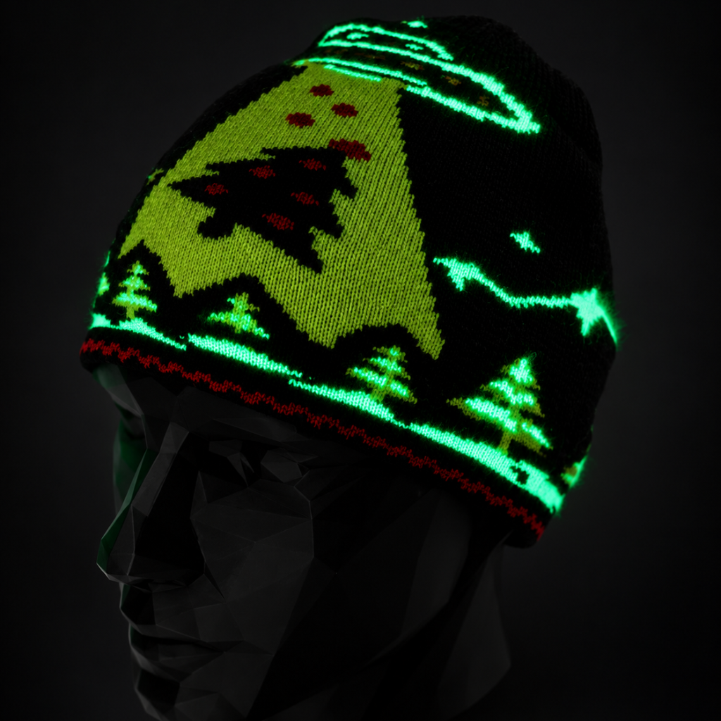 A black knit beanie with a glowing green Christmas tree and mountain pattern, displayed on a black mannequin head against a dark background.