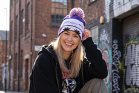 Person wearing a black hoodie and colorful beanie squatting on a street with graffiti in the background
