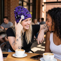 Two women sitting at a table with coffee, one wearing a purple beanie with haunted mansion wallpaper print.