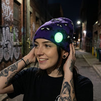 Person wearing a haunted mansion beanie with a green light on a graffiti-covered street at night.