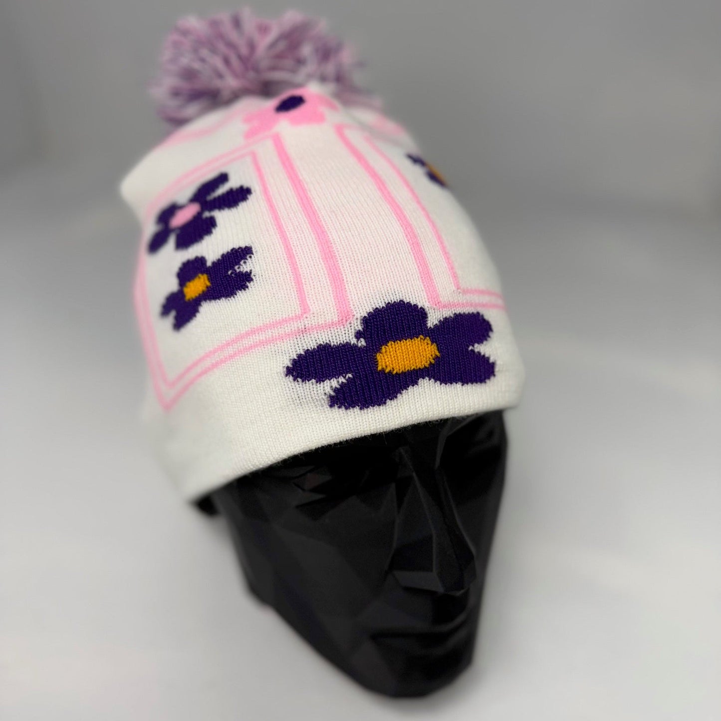 White knit hat with purple flowers and a pom-pom on a mannequin head against a gray background