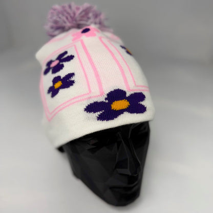 White knit hat with purple flowers and a pom-pom on a mannequin head against a gray background