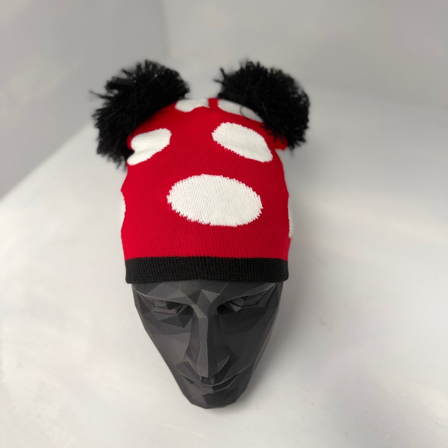 Red and white polka dot hat with black pom-poms on a mannequin head against a gray background