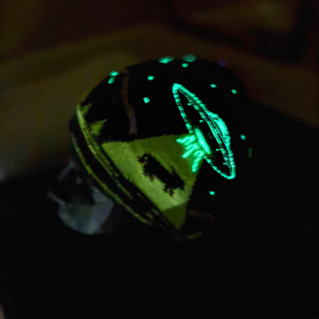 A black knit beanie with glowing green and yellow glow-in-the-dark graphics, shown in a dark environment to highlight its luminescent properties.