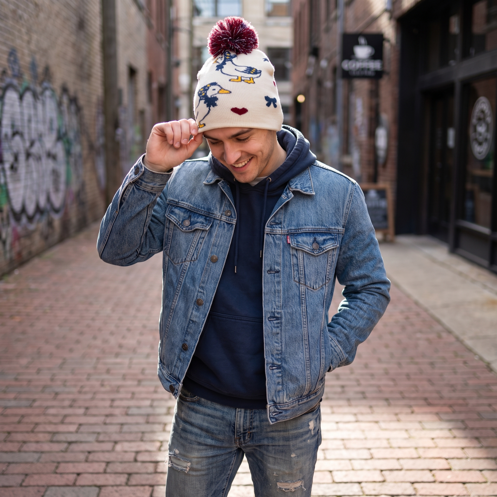 A man wearing a light blue denim jacket, navy hoodie, and a cream knit beanie with a maroon pom-pom and colorful duck illustrations, standing in a brick alleyway.