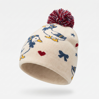 An off-white knit pom-pom beanie featuring a pattern of blue ducks, yellow beaks, red hearts, and blue bows, topped with a maroon and white pom-pom, shown against a light gray background.