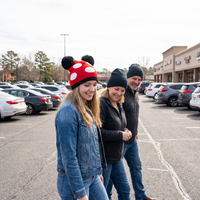Three people walking in a parking lot with a person wearing a red and white hat with ears.