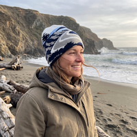 A woman smiling on a beach wearing a navy, white, and gray patterned knit beanie and a tan hooded jacket, with ocean waves and rocky cliffs in the background.