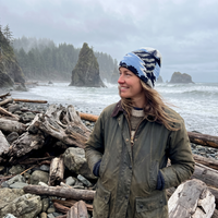 A woman standing on a rocky beach with driftwood logs in the foreground and sea stacks under a cloudy sky in the background. She is wearing an olive green jacket and a blue and white patterned knit beanie.