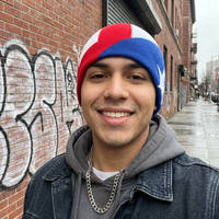 Man wearing a red, white, and blue beanie in front of a brick wall with graffiti.