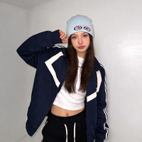 Person wearing a navy blue jacket with white accents, white beanie, and black pants against a plain background