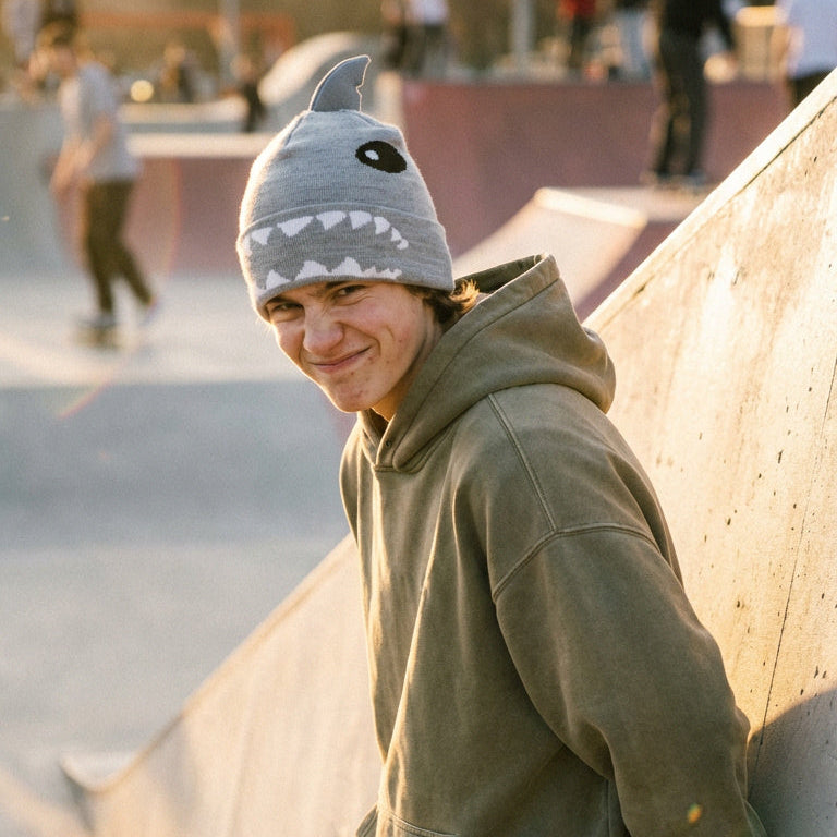 Person wearing a shark-themed beanie and green hoodie leaning against a concrete wall at a skate park.