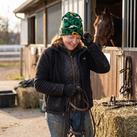 Person standing next to a horse in a stable area, holding reins, weating a celtic beanie