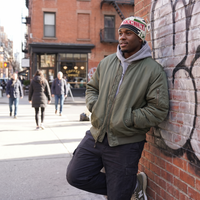 A man wearing an olive green bomber jacket, black cargo pants, a gray hoodie, and a colorful knit beanie leans against a brick wall on a city street.