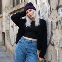 Person wearing a black beanie with 'CRAZY' text, black crop top, and blue jeans against a graffiti-covered wall.