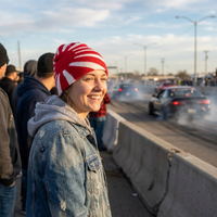 Person wearing a red and white beanie with a pattern, standing on a bridge with cars and people in the background.