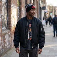 Man wearing a black bomber jacket and beanie standing on a street with graffiti on the wall.
