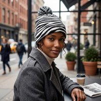 Woman wearing a striped beanie and gray coat sitting outdoors with a newspaper and coffee.