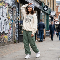 Woman in a beige sweatshirt and green pants standing on a street with graffiti and a coffee shop in the background.