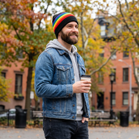 Man wearing a rainbow beanie and denim jacket holding a coffee cup in an urban setting with trees and buildings.