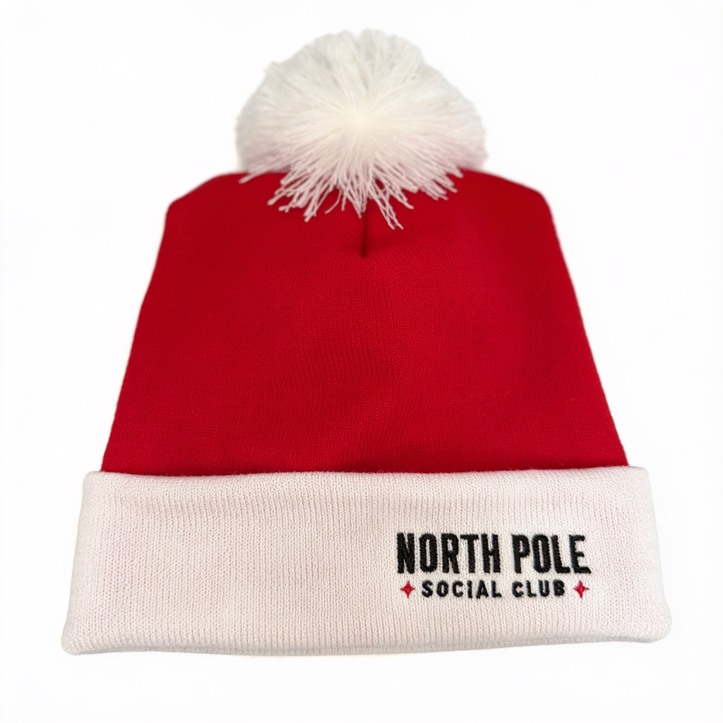 A red knit Santa hat with a white pom-pom on top and a white ribbed cuff. The cuff features the embroidered text 'NORTH POLE SOCIAL CLUB' in black with small red star accents.
