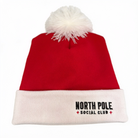A red knit Santa hat with a white pom-pom on top and a white ribbed cuff. The cuff features the embroidered text 'NORTH POLE SOCIAL CLUB' in black with small red star accents.