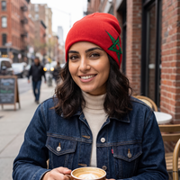 A woman wearing a bright red knit beanie with a green embroidered star, a dark blue denim jacket, and a cream turtleneck, holding a cup of coffee on a city street.