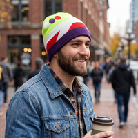 A smiling man wearing a colorful knit beanie with green, white, red, and purple sections, a denim jacket, and holding a takeaway coffee cup on a city street.