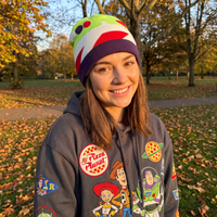 A smiling young woman outdoors in a park wearing a gray hoodie with multiple Buzz Lightyear and Toy Story patches, along with a colorful knit beanie.