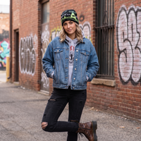 A woman stands in an urban alley wearing a light-wash blue denim jacket over a gray graphic hoodie, black ripped jeans, and brown lace-up boots. She is also wearing a black beanie with green and white lettering.