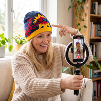 A woman wearing a colorful knit beanie sits on a couch indoors, holding a phone in front of a ring light. The beanie has a navy blue top with red and yellow graphic patterns and a yellow brim.