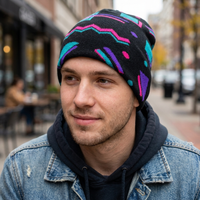 A man wearing a black knit beanie with a colorful geometric pattern in pink, purple, and teal, styled with a dark hoodie and a distressed denim jacket in an outdoor urban setting.